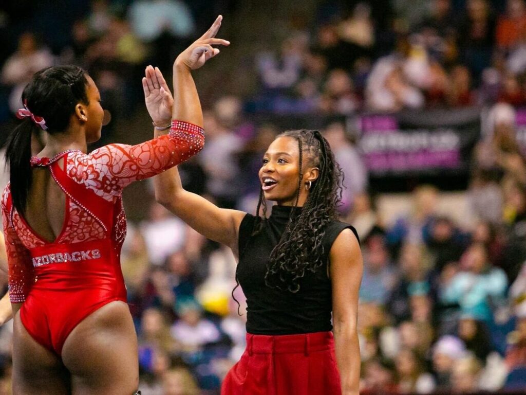 Two gymnasts celebrate with a high-five on the competition floor; one wears a red patterned leotard, the other a black sleeveless top with a red skirt.