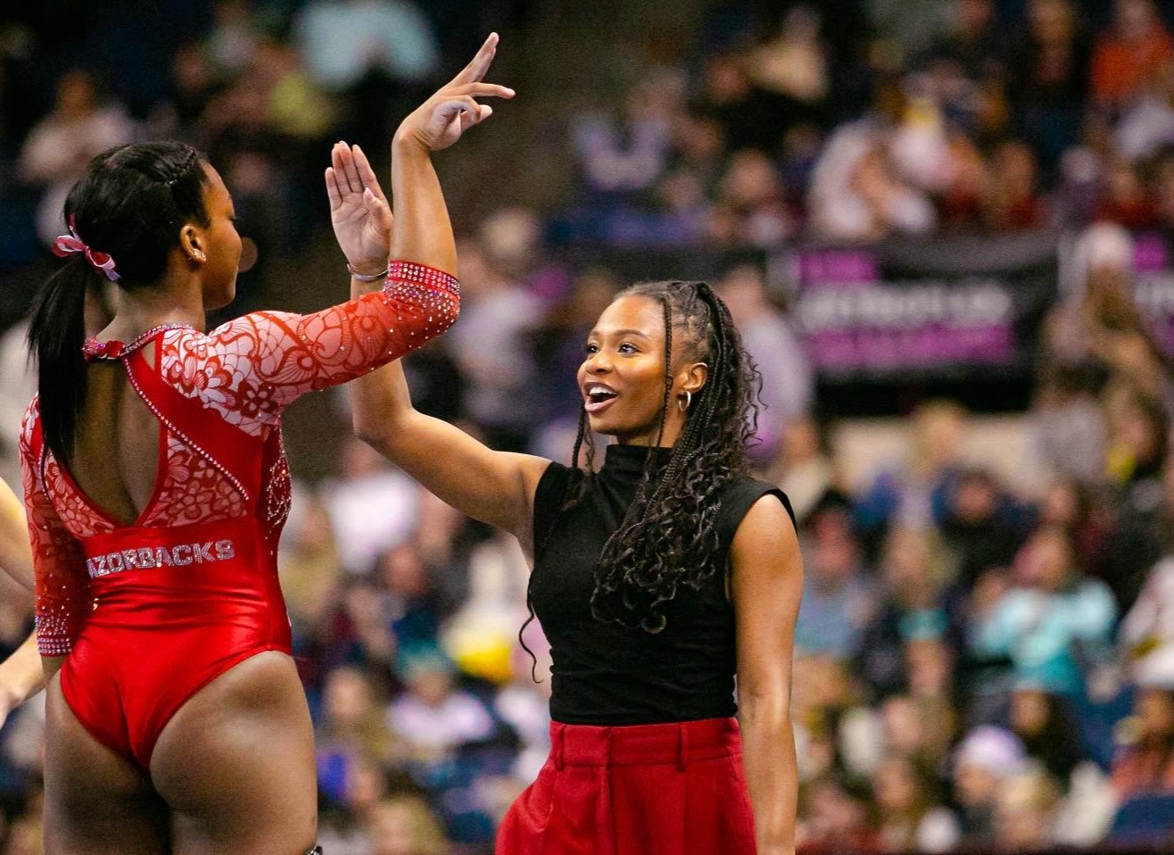 Two gymnasts celebrate with a high-five on the competition floor; one wears a red patterned leotard, the other a black sleeveless top with a red skirt.