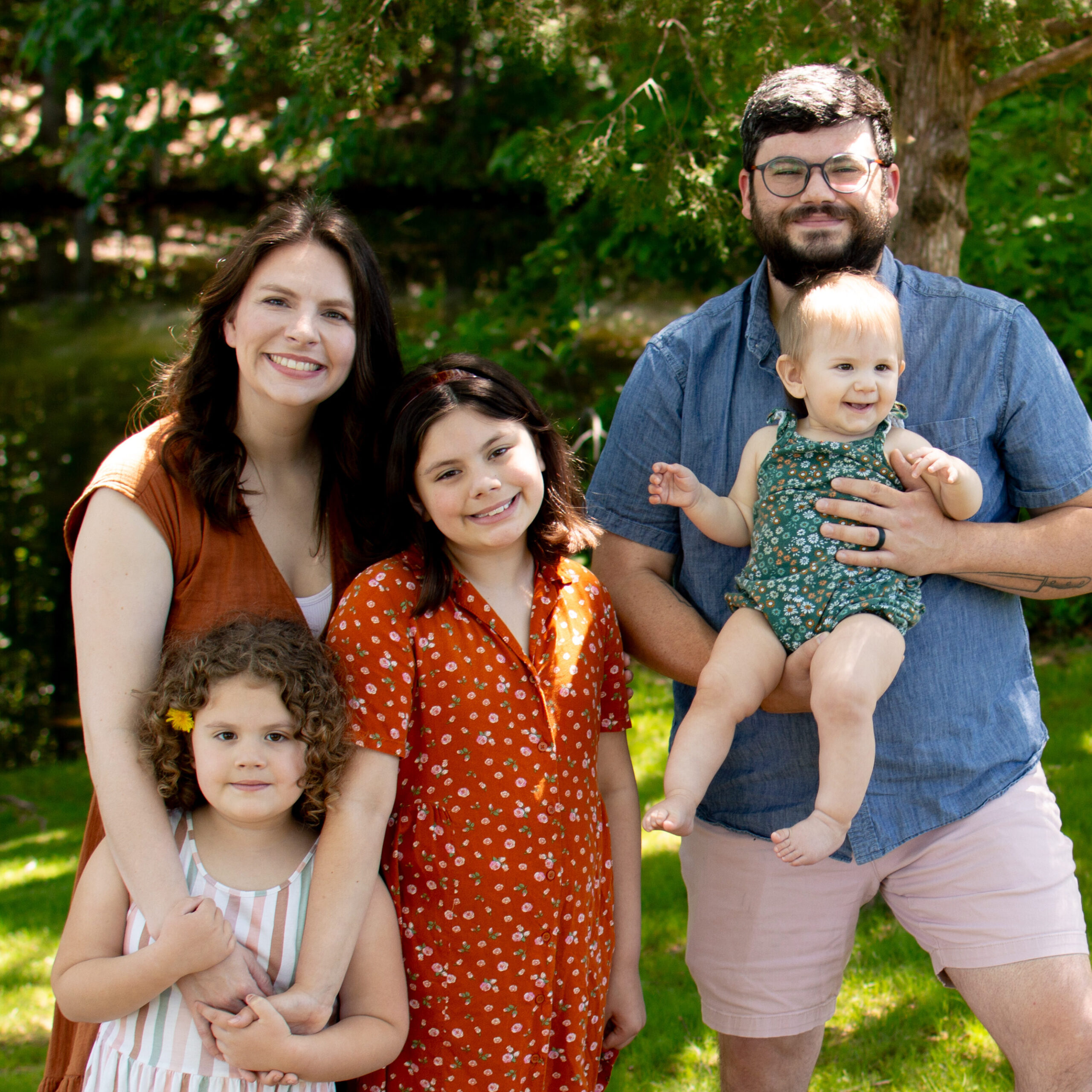 A family of six posing outdoors by a grassy area with a tree, smiling at the camera (two women, two girls, a man, and a baby).